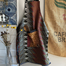 Load image into Gallery viewer, Handcrafted African wax print apron made in Uganda. Perfect for housewarming and wedding gitfs. Excellent for entertaining guests during the holidays, Thankgiving, Christmas, and more. Shown with olivewood salad server.
