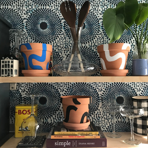 Handpainted terracotta planters with Bauhaus bohemian painted designed. Perfect housewarming gift for herbs, succulents, and indoor house plants. African textile wallpaper in the background with open shelving and Ugandan salad servers.