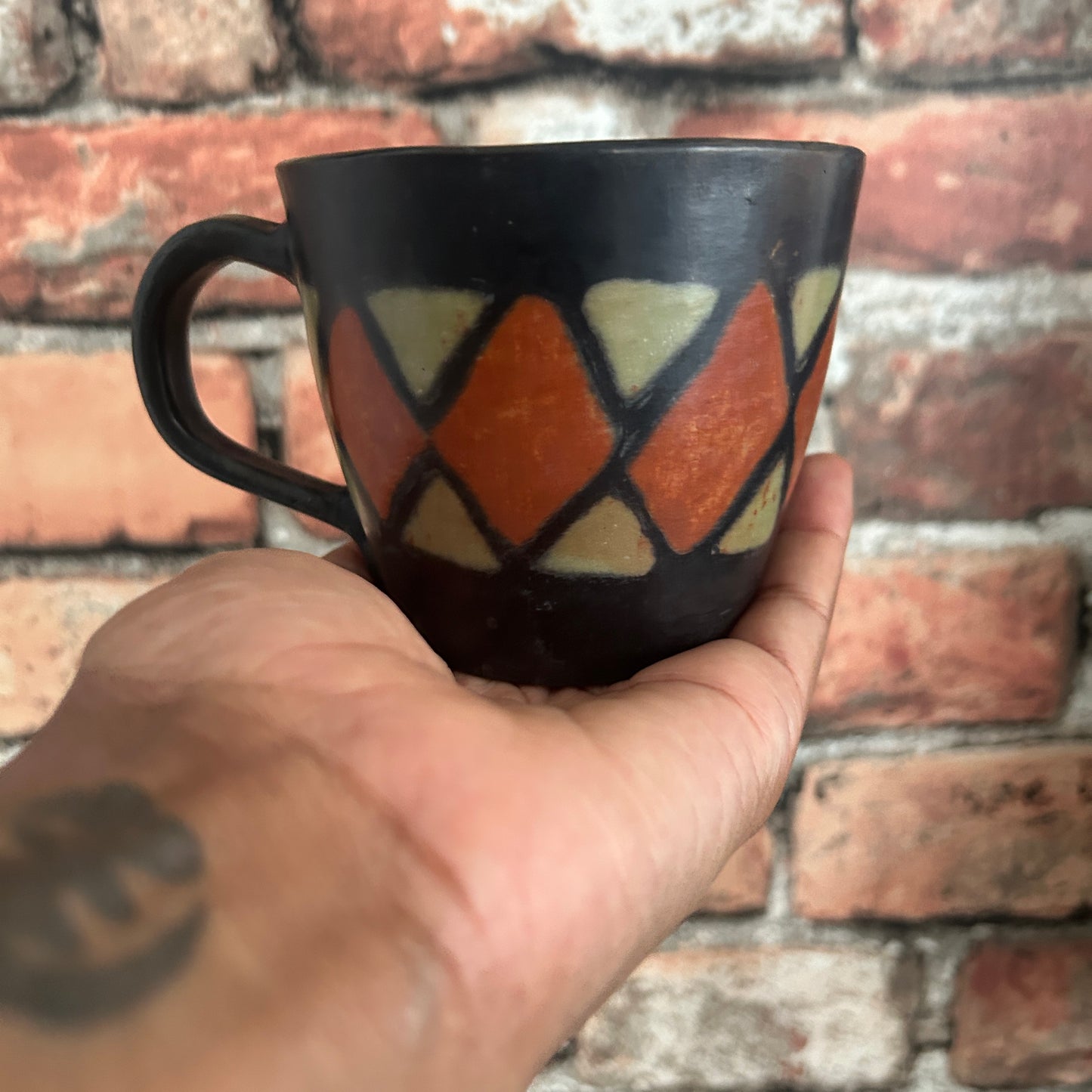 Hand holding a black mug with geometric pattern against a brick wall