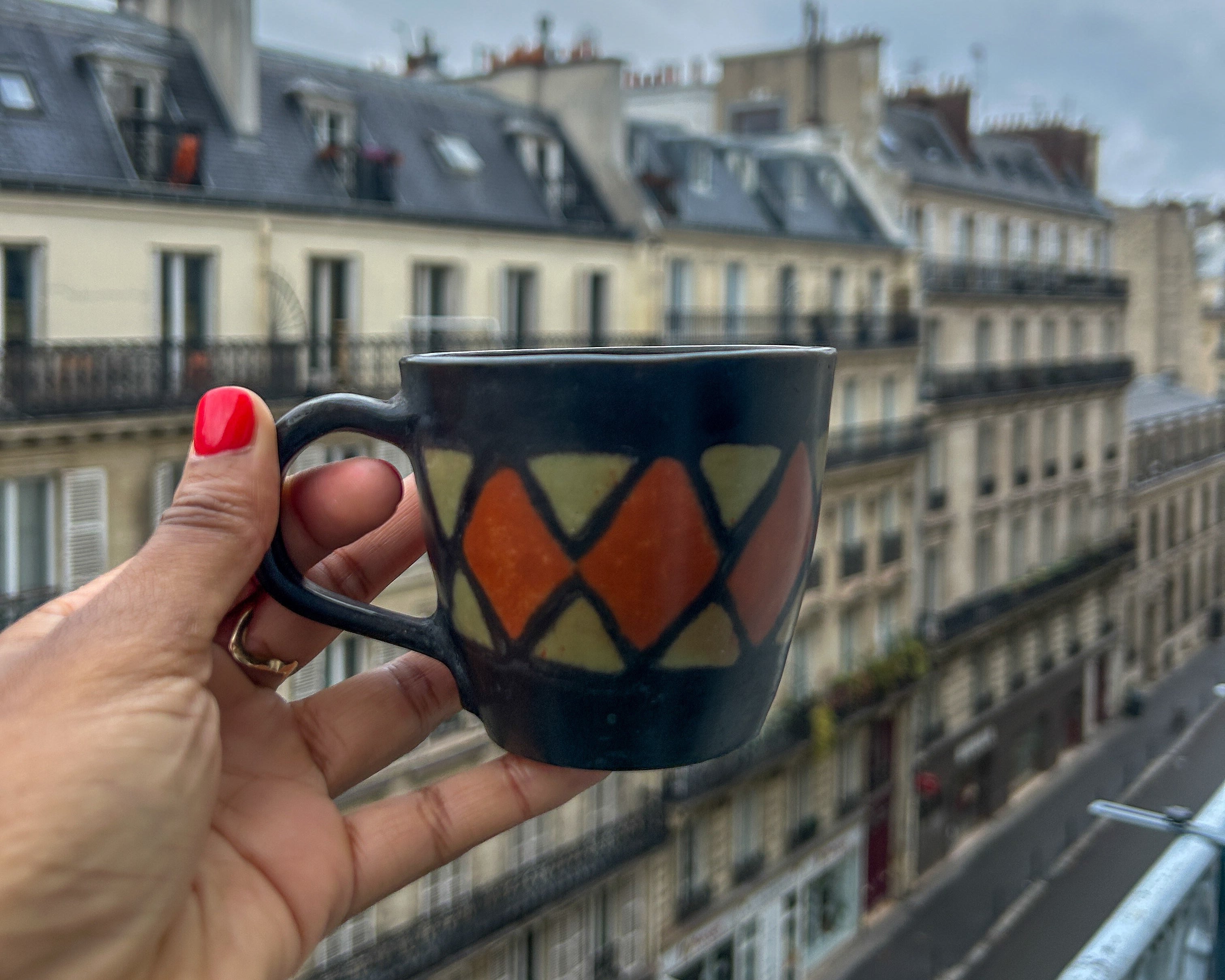 Person holding a mug with a geometric design, looking out over a city street with buildings in the background.