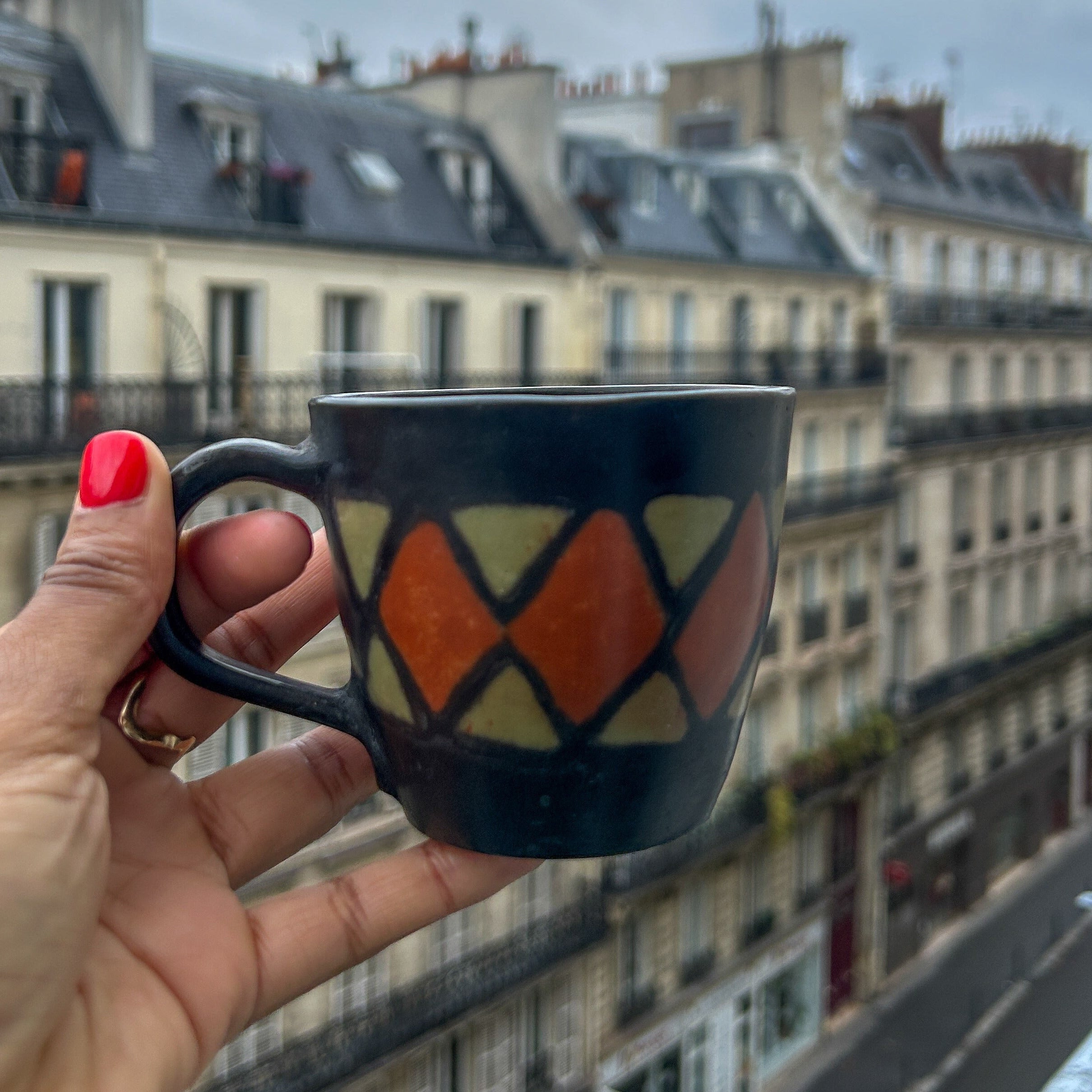 Person holding a mug with a geometric design, looking out over a Paris city street with buildings in the background.