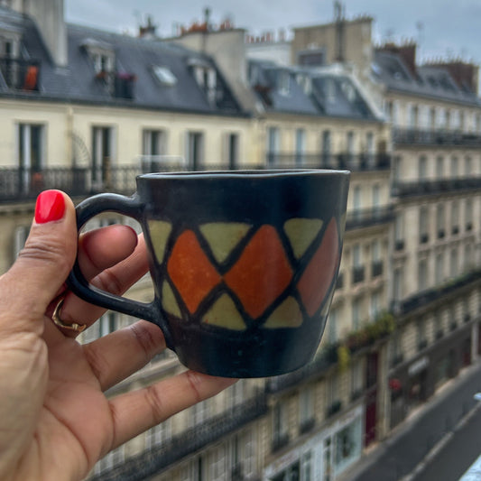 Person holding a mug with a geometric design, looking out over a Paris city street with buildings in the background.