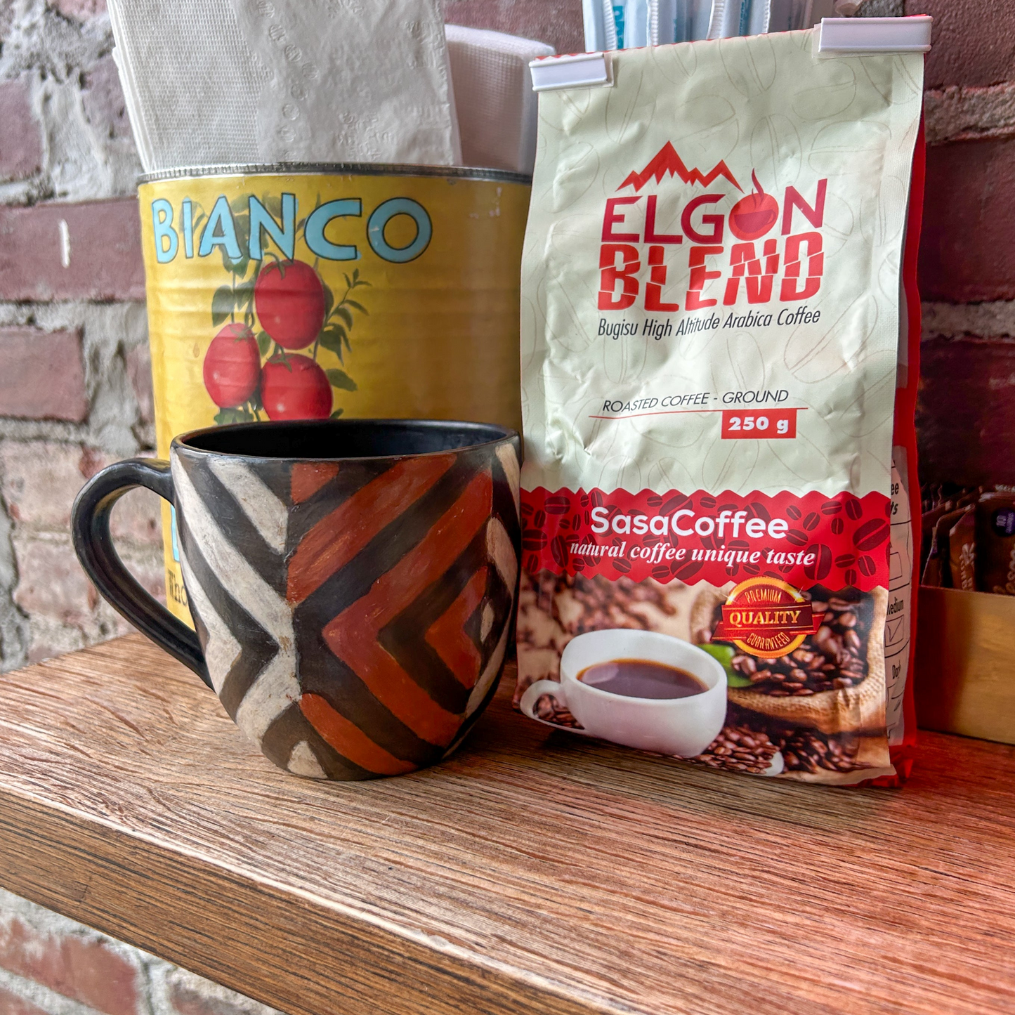 Coffee mug and bags of Ugandan coffee on a wooden surface with a brick wall background in a Brooklyn cafe.