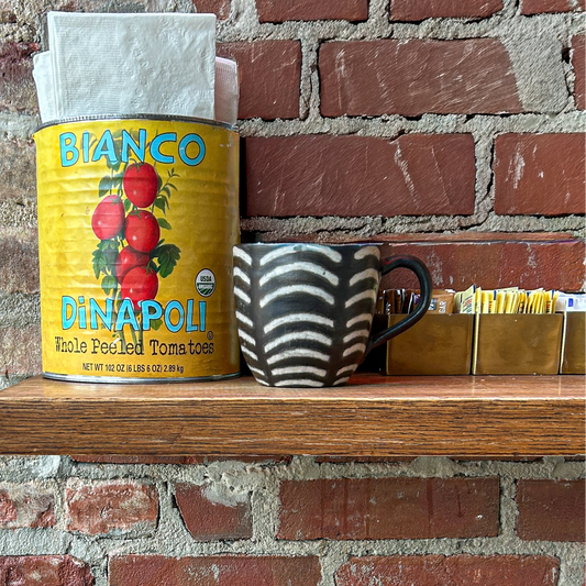 Shelf with a can of Bianco D'Anapo, a Ugandan mug against a brick wall in a Brooklyn cafe.