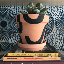 Load image into Gallery viewer, Handpainted terracotta planters with Bauhaus bohemian painted designed. Seen here in black. Perfect housewarming gift for herbs, succulents, and indoor house plants. African textile wallpaper in the background with open shelving.
