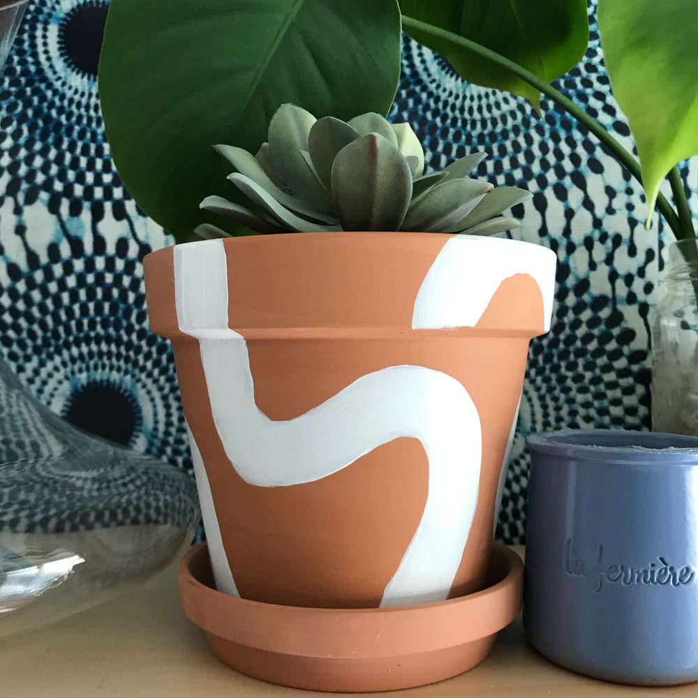 Handpainted terracotta planters with Bauhaus bohemian painted designed in white. Perfect housewarming gift for herbs, succulents, and indoor house plants. African textile wallpaper in the background with open shelving.