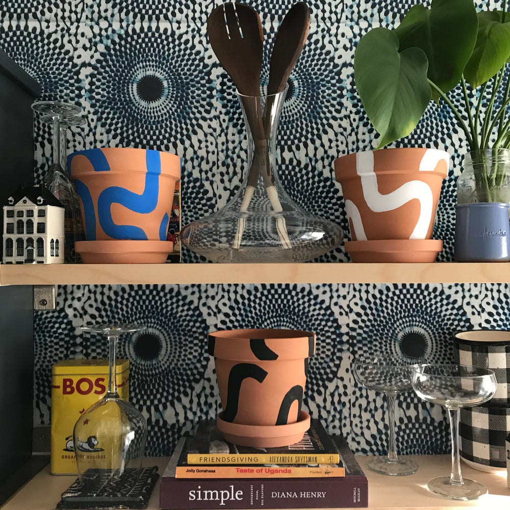 Handpainted terracotta planters with Bauhaus bohemian painted designed. Perfect housewarming gift for herbs, succulents, and indoor house plants. African textile wallpaper in the background with open shelving and Ugandan salad servers.