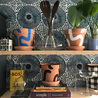 Handpainted terracotta planters with Bauhaus bohemian painted designed. Perfect housewarming gift for herbs, succulents, and indoor house plants. African textile wallpaper in the background with open shelving and Ugandan salad servers.