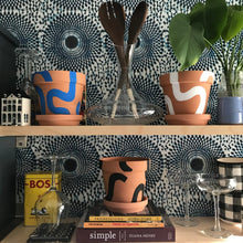 Load image into Gallery viewer, Handpainted terracotta planters with Bauhaus bohemian painted designed. Perfect housewarming gift for herbs, succulents, and indoor house plants. African textile wallpaper in the background with open shelving and Ugandan salad servers.
